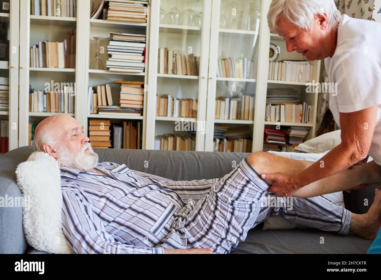 Carer tratta l'osteoartrite del ginocchio senior sul divano a casa con fisioterapia Foto Stock