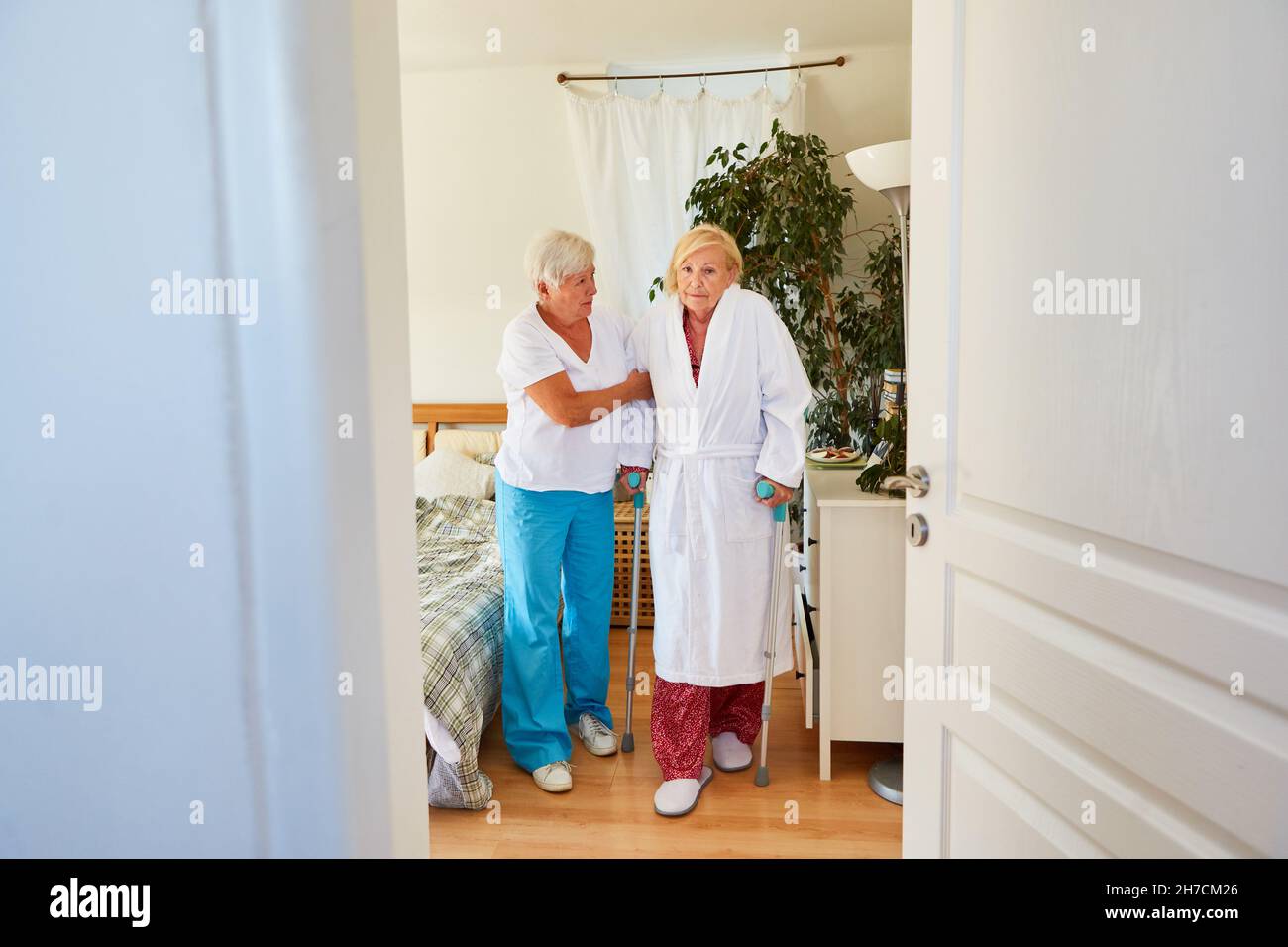 L'operatore sanitario aiuta la donna anziana in un accappatoio con stampelle mentre cammina nella clinica di riabilitazione o a casa Foto Stock