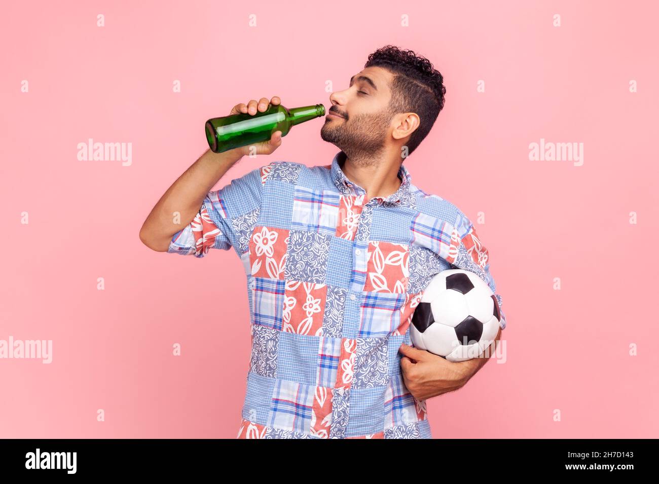 Vista laterale dell'uomo brunetto con barba che indossa una camicia blu casual in cui tieni la palla di calcio in mano e beve alcol mentre rallegrano la squadra preferita. Studio interno girato isolato su sfondo rosa. Foto Stock