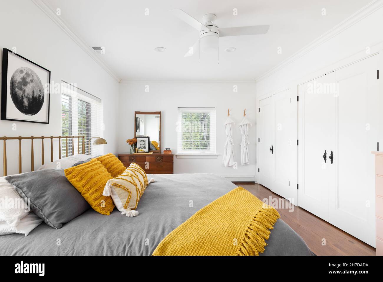 Una luminosa camera da letto con pareti bianche, biancheria da letto gialla e grigia e un tappeto su pavimenti in legno. Foto Stock