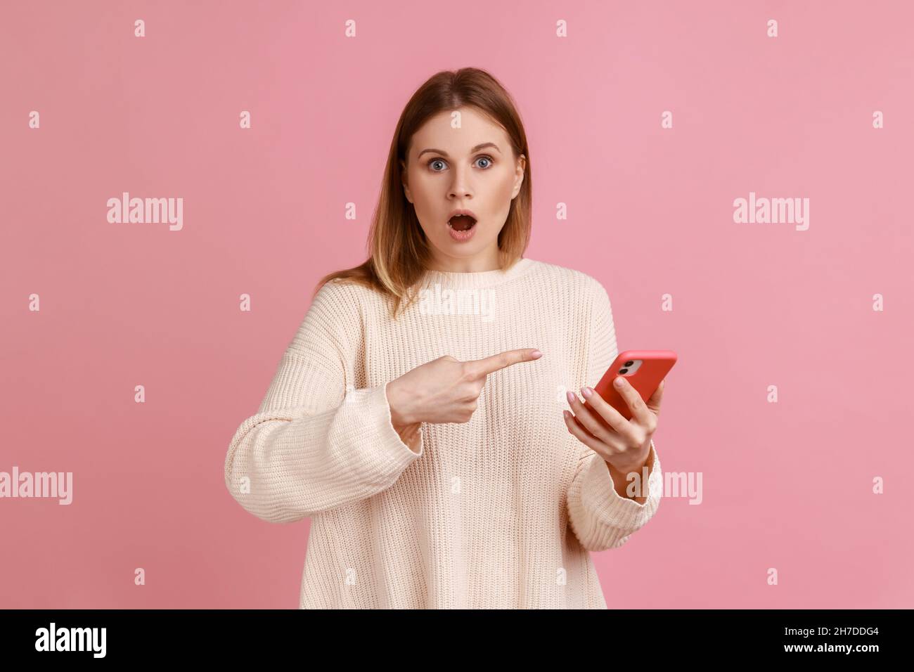 Ritratto di stupito bionda donna blogger tenere il telefono cellulare in mani, puntando al gadget con stupore e bocca aperta, indossando maglione bianco. Studio interno girato isolato su sfondo rosa. Foto Stock