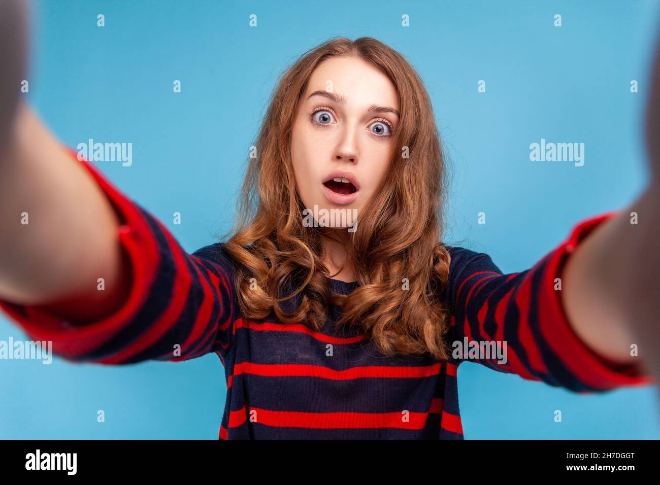 Donna sorpresa che indossa un pullover casual a righe, con selfie dal volto scioccato, con occhi grandi e bocca aperta, POV. Studio interno girato isolato su sfondo blu. Foto Stock