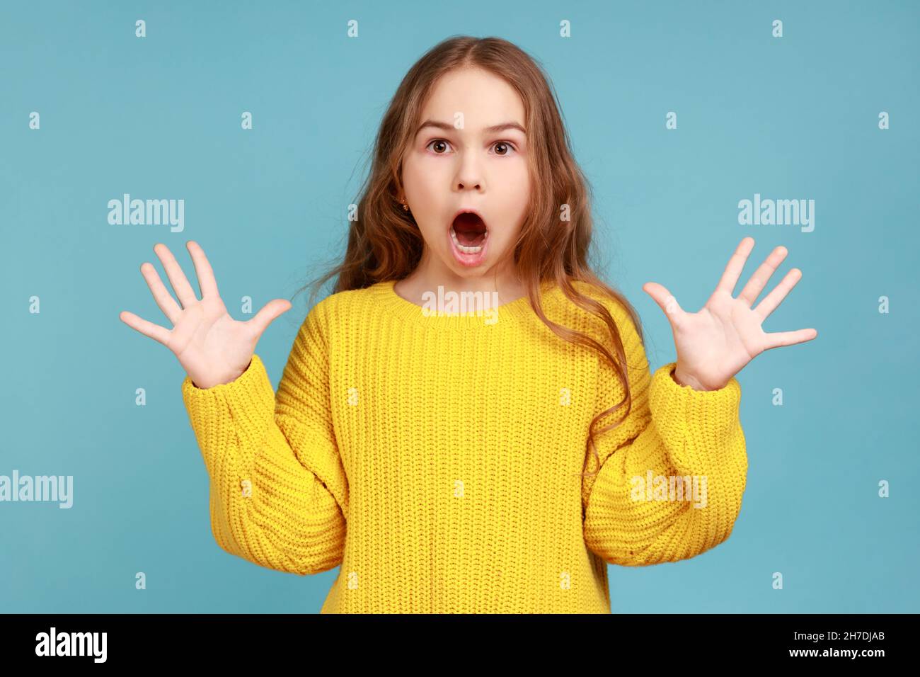 Ritratto di spaventato scioccata bambina in piedi con le braccia sollevate e guardando la macchina fotografica con gli occhi grandi, indossando un maglione giallo casual stile. Studio interno girato isolato su sfondo blu. Foto Stock