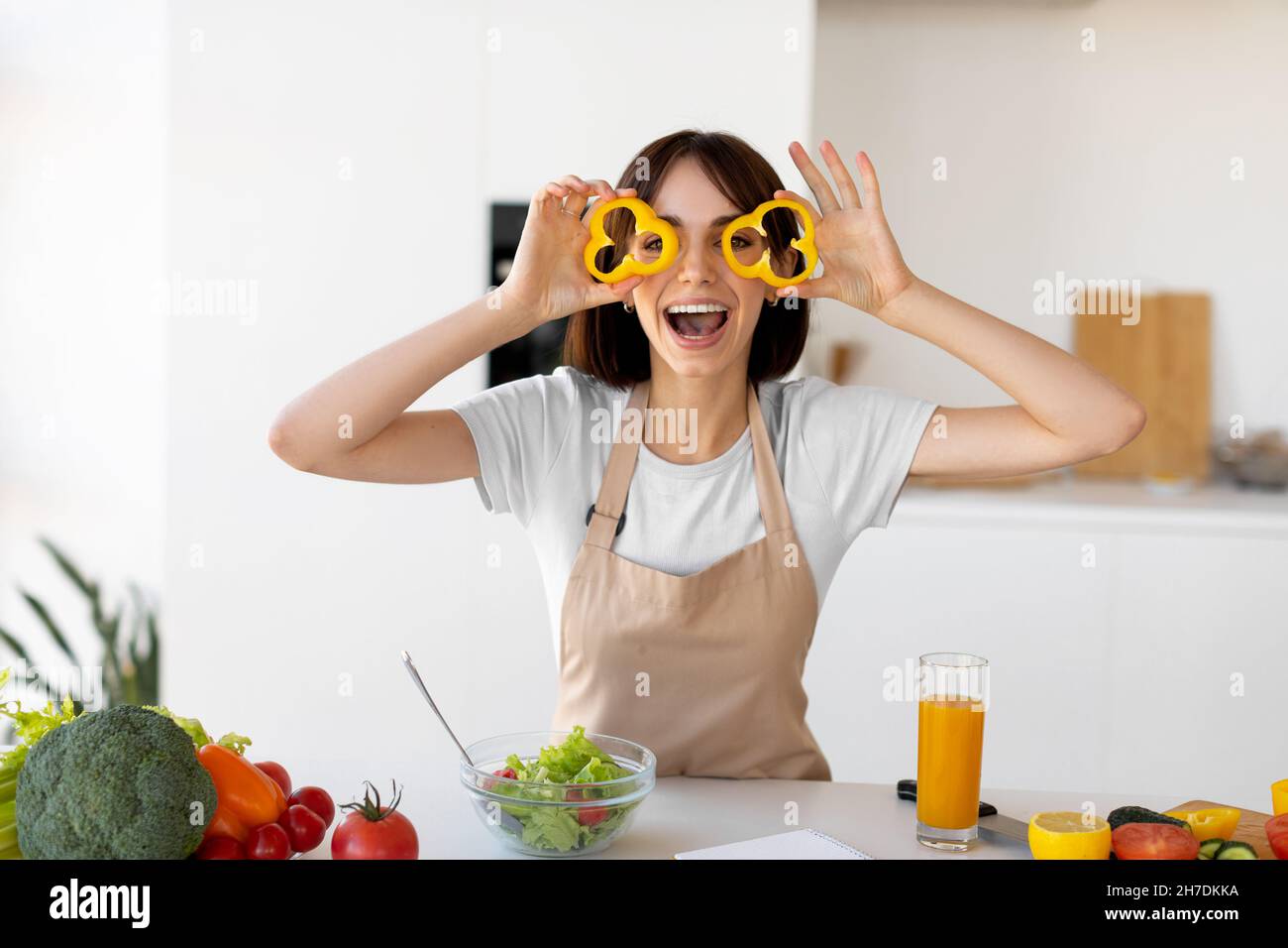 Gioiosa giovane donna che fa bicchieri di peperone dolce, divertirsi e giocare con il cibo mentre cucinano insalata fresca in cucina Foto Stock