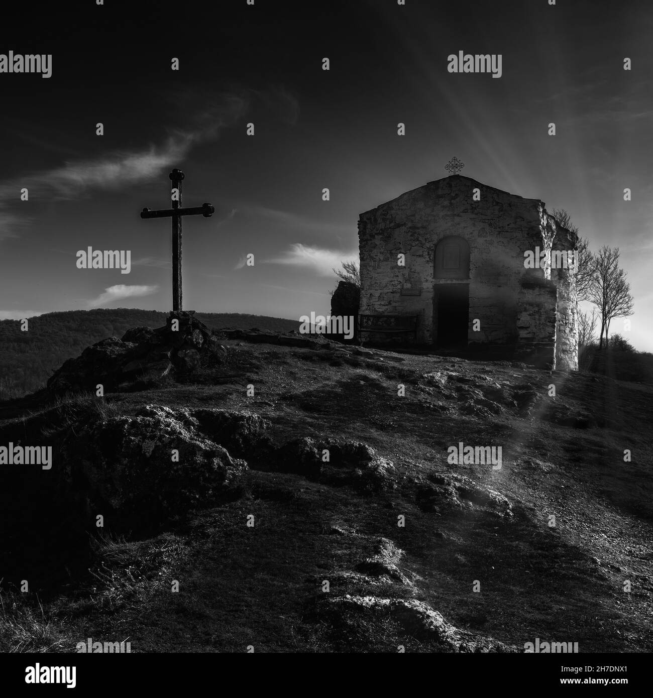 Foto in bianco e nero di una piccola cappella medievale ortodossa e croce sulla cima di una collina rocciosa. Retroilluminazione dei raggi solari Foto Stock