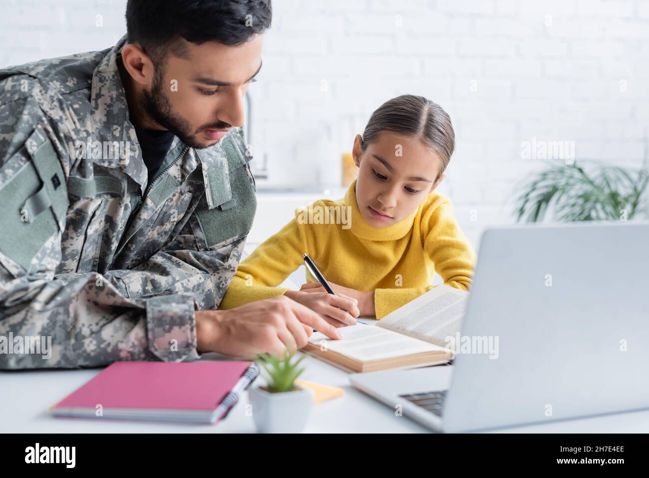 Padre in uniforme militare che indica il libro vicino alla figlia e computer portatile a casa Foto Stock