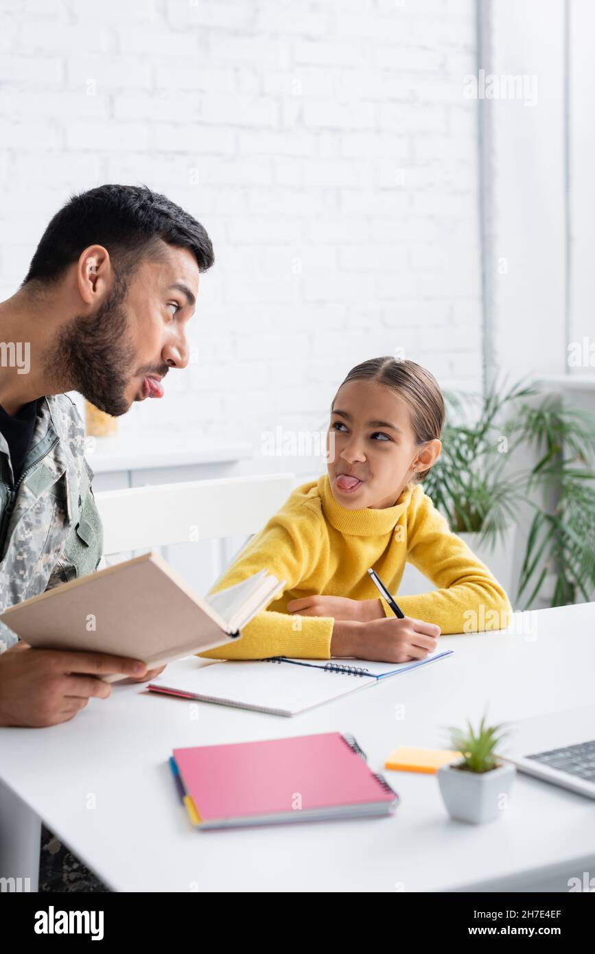 Uomo in uniforme militare che tiene il libro vicino alla figlia che attacca fuori la linguetta e scrive sul taccuino nel paese Foto Stock
