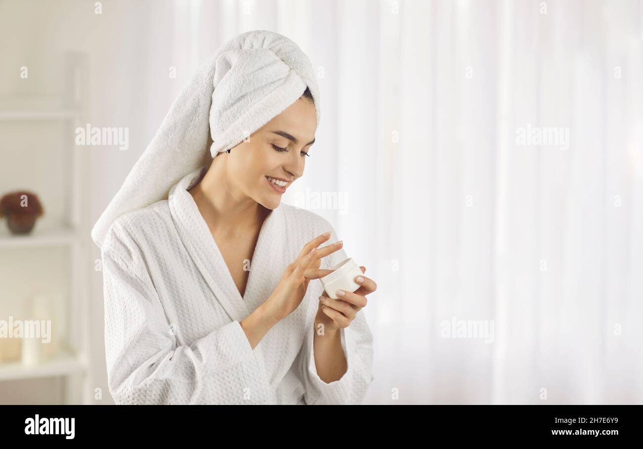 Giovane donna in accappatoio e asciugamano che fa la routine di cura della pelle e applicazione di crema per il viso Foto Stock