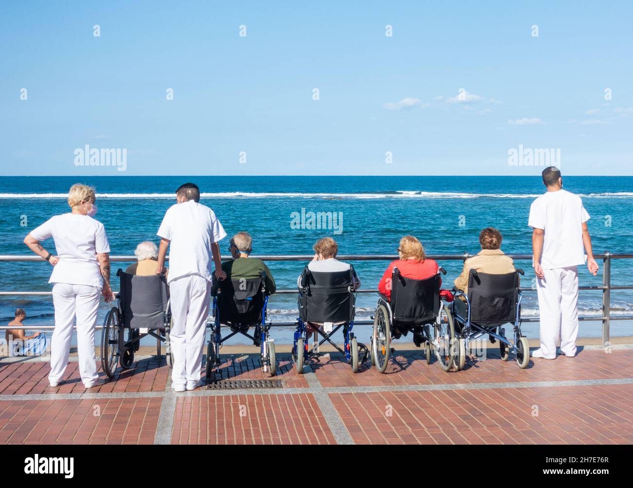 Anziani assistenza a casa residenti in carrozzina guardando al mare in Spagna con accompagnatori in uniformi bianche Foto Stock