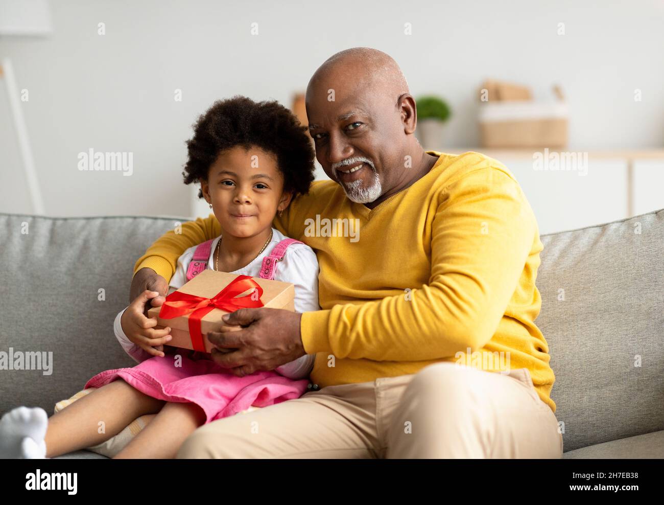 Sorridendo uomo anziano nero dà il regalo di compleanno, bambino piccolo apre la scatola nell'interno del soggiorno Foto Stock