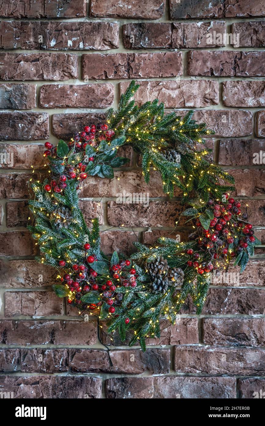 Una bella corona di Natale isolato su un muro di mattoni sfondo. Corona di Natale fatta a mano fatta di abete, bacche rosse e coni di pino appesi al muro per il saluto della stagione delle feste. Scatto verticale Foto Stock