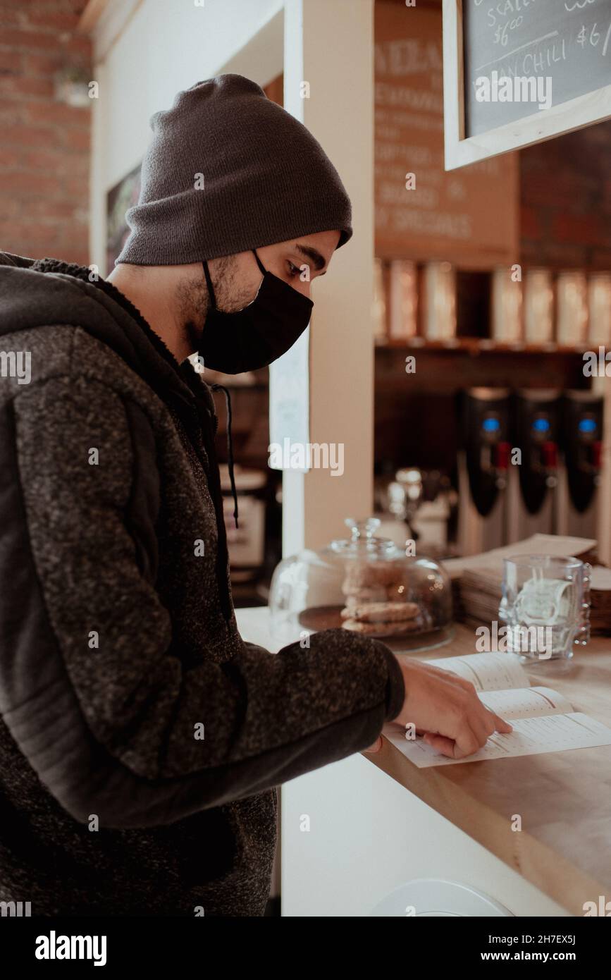 Misto-razza giovane uomo ordini al banco di quartiere caffè indossando una maschera nera e beanie. In bar indossare maschera e sociale distanziamento durante COVID Foto Stock