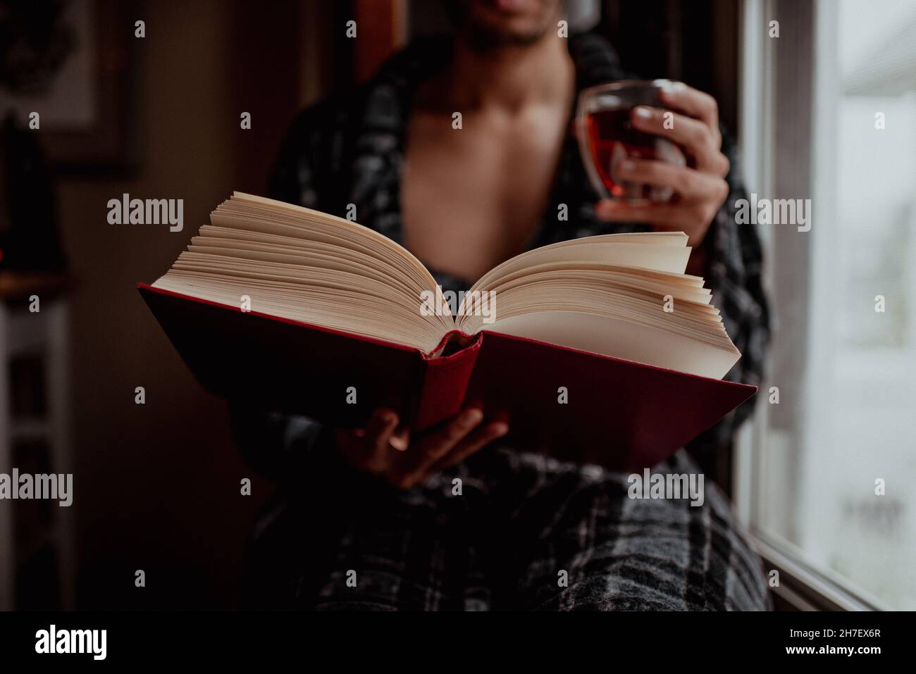 Il giovane uomo di razza mista siede sul sedile della finestra durante la lettura nuvolosa di giorno e bevendo il tè o il caffè. Cura di sé e me-tempo a casa. Relax e relax. Foto Stock