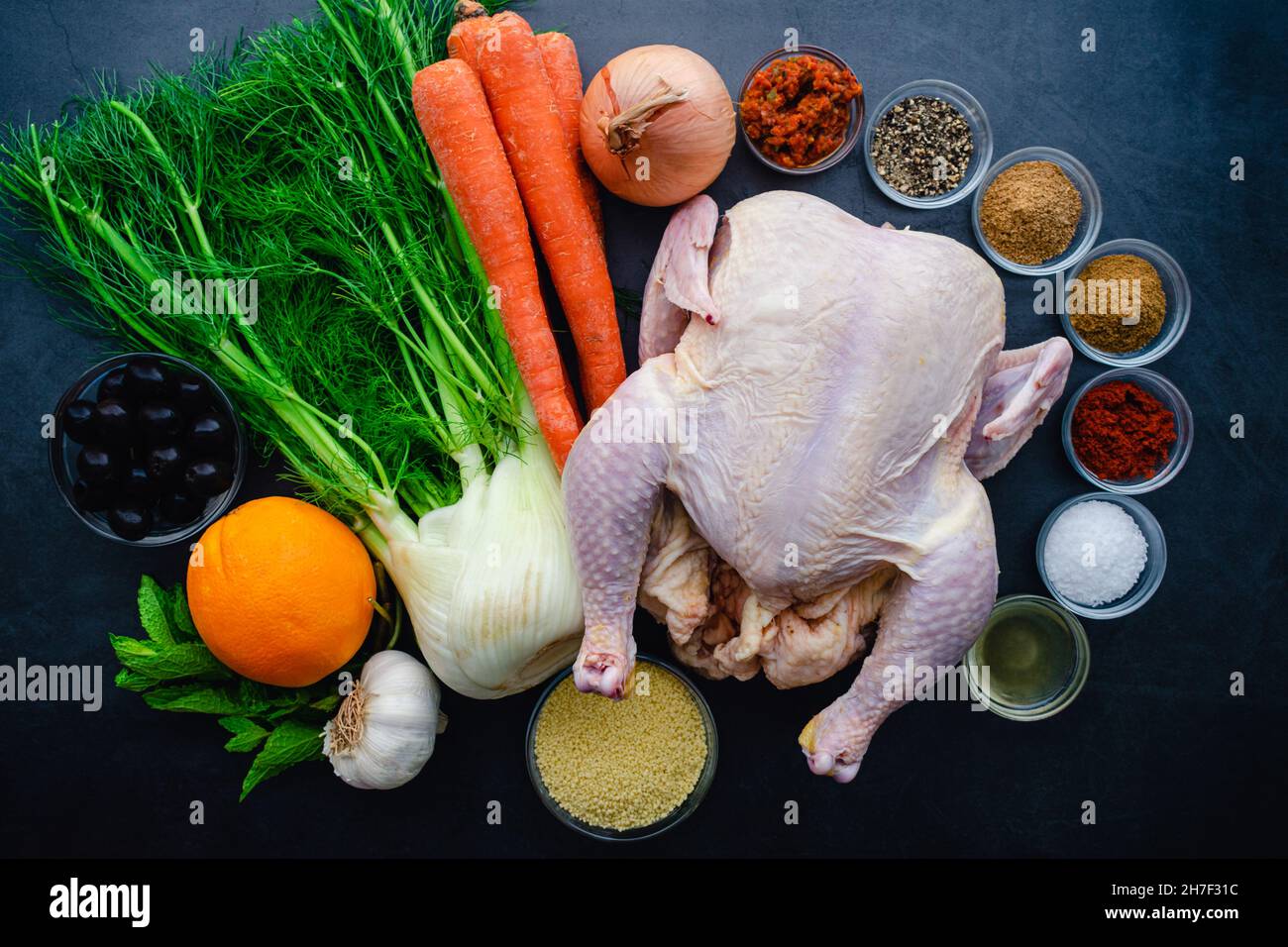 Ingredienti per il pollo speziato marocchino con verdure arrosto: Vista dall'alto di pollo crudo, verdure e spezie su sfondo scuro Foto Stock