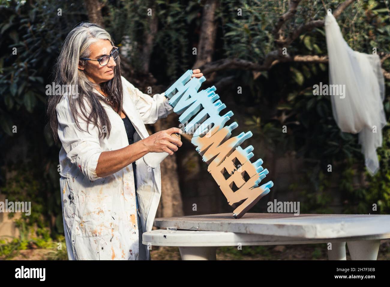 Donna con capelli grigi che dipinge alcune lettere di polistirene nel giardino Foto Stock