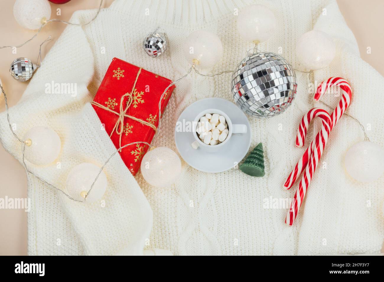 Tazza di caffè, scatola regalo e maglioni in maglia, luci a LED su sfondo beige. Stile Higge, concetto invernale. Accogliente scrivania. Capodanno e Natale Foto Stock