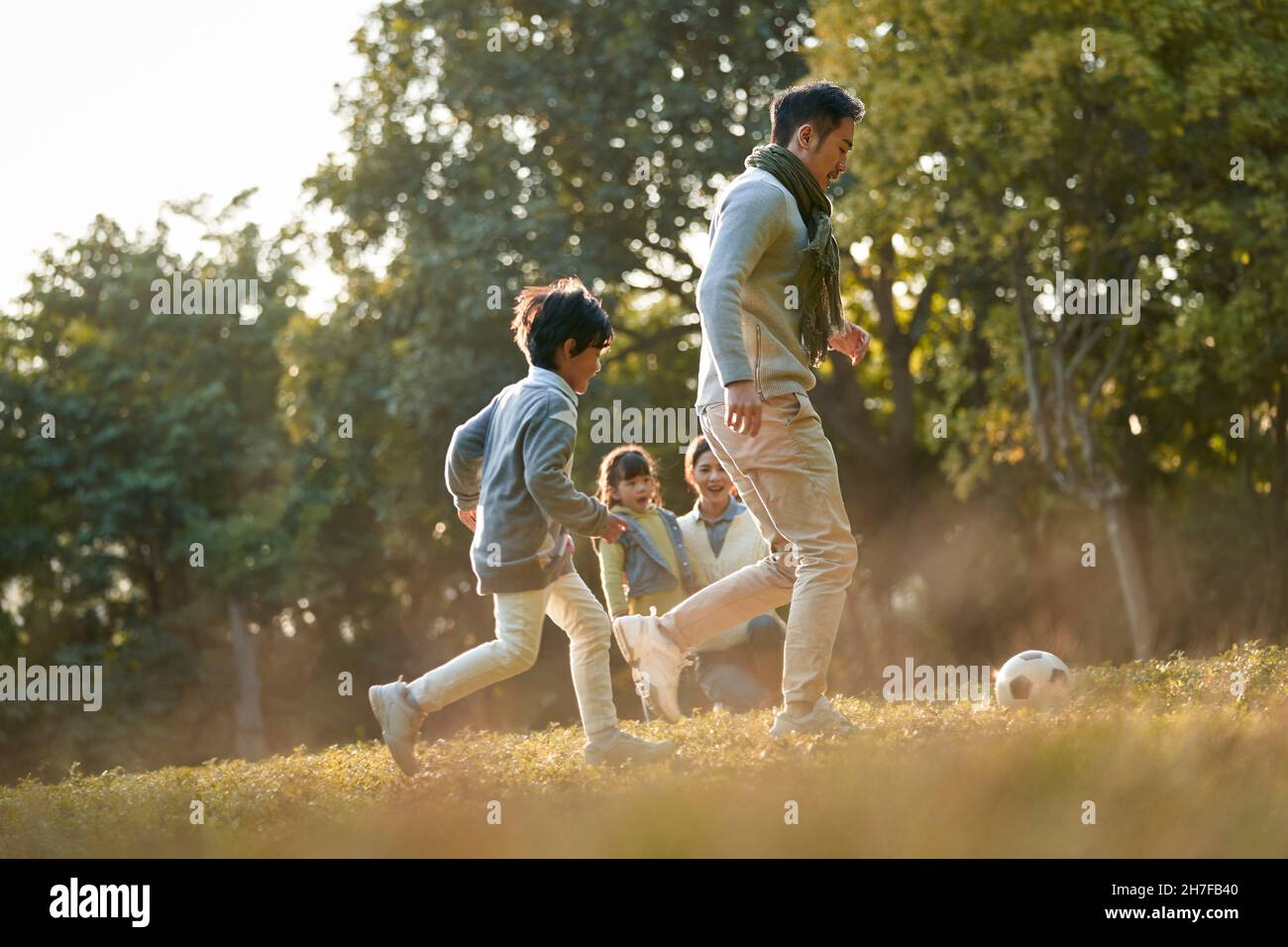padre e figlio asiatici che giocano a calcio all'aperto nel parco, mentre madre e figlia guardano da dietro Foto Stock