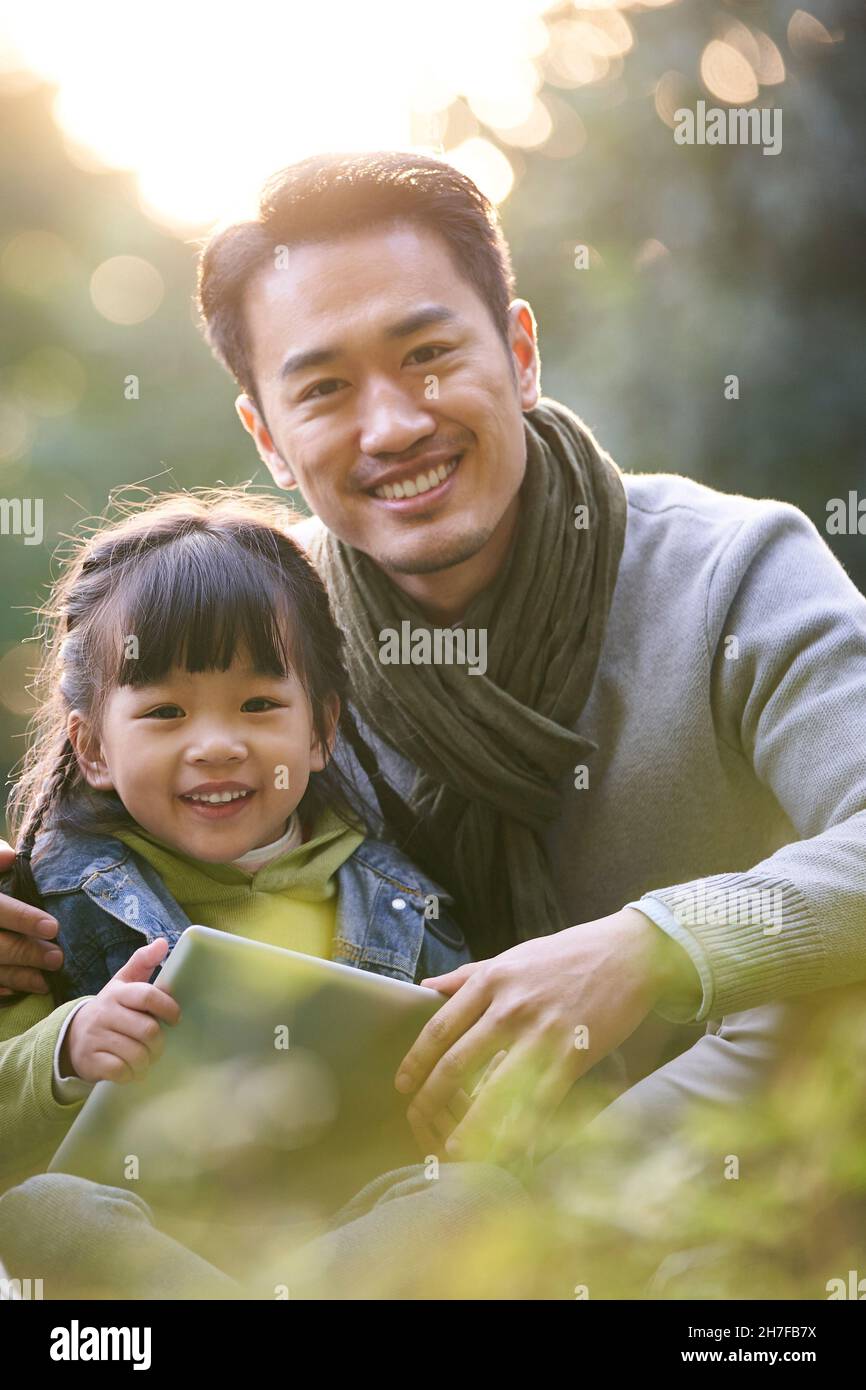 ritratto all'aperto di un padre asiatico e di una figlia seduta sull'erba guardando la macchina fotografica sorridente Foto Stock