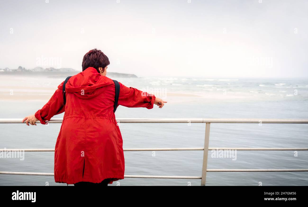 Vista posteriore di una donna riflettente trench rosso che contempla il mare su una spiaggia in una vacanza estiva, all'aperto. Stile di vita di viaggio di mezza età. Foto Stock