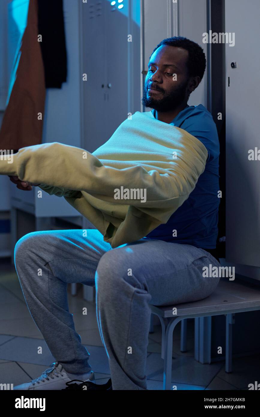 Sportivo africano con portici in t-shirt e pantaloni sudoripani indossando una felpa mentre si siede da una fila di armadietti in spogliatoio Foto Stock