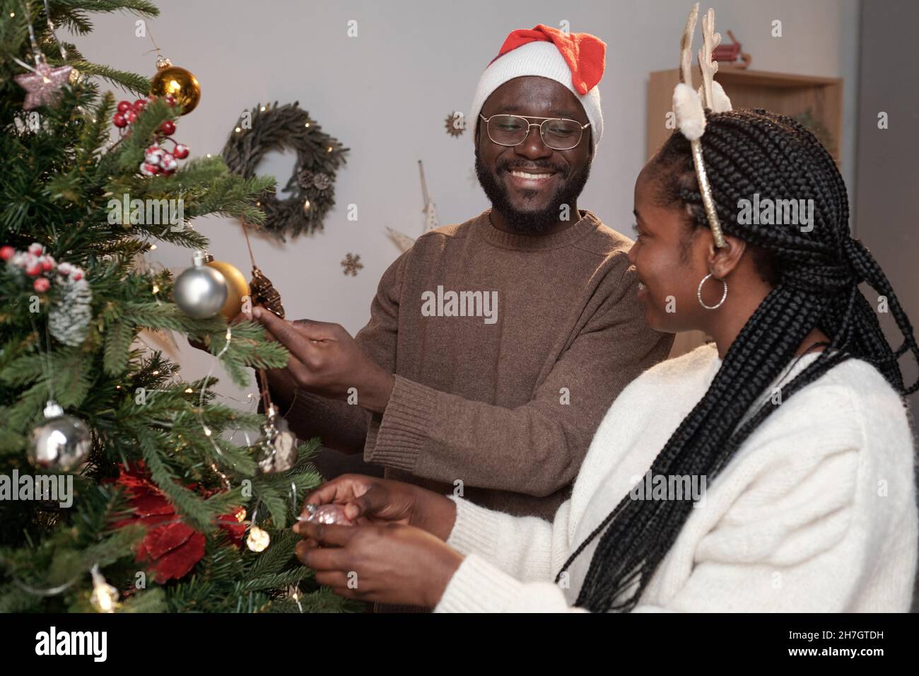 Giovane uomo africano sorridente in occhiali, maglione e Babbo Natale a guardare sua moglie, mentre entrambi decorare albero di Natale con giocattoli e palle Foto Stock