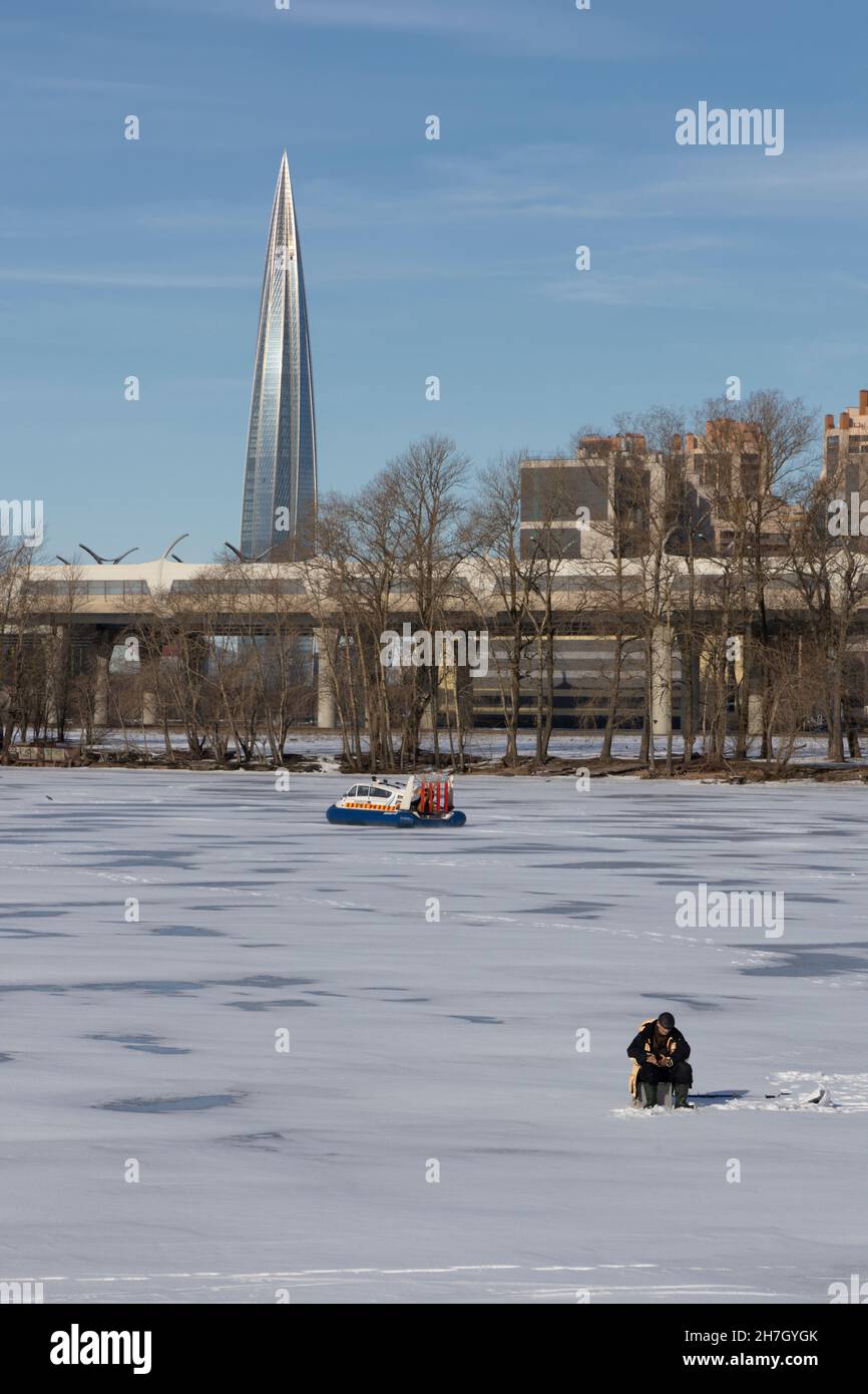San Pietroburgo, Russia - 24 marzo 2021: L'uomo è impegnato nella pesca invernale sul ghiaccio del fiume e un hovercraft EMERCOM passa vicino contro il bac Foto Stock
