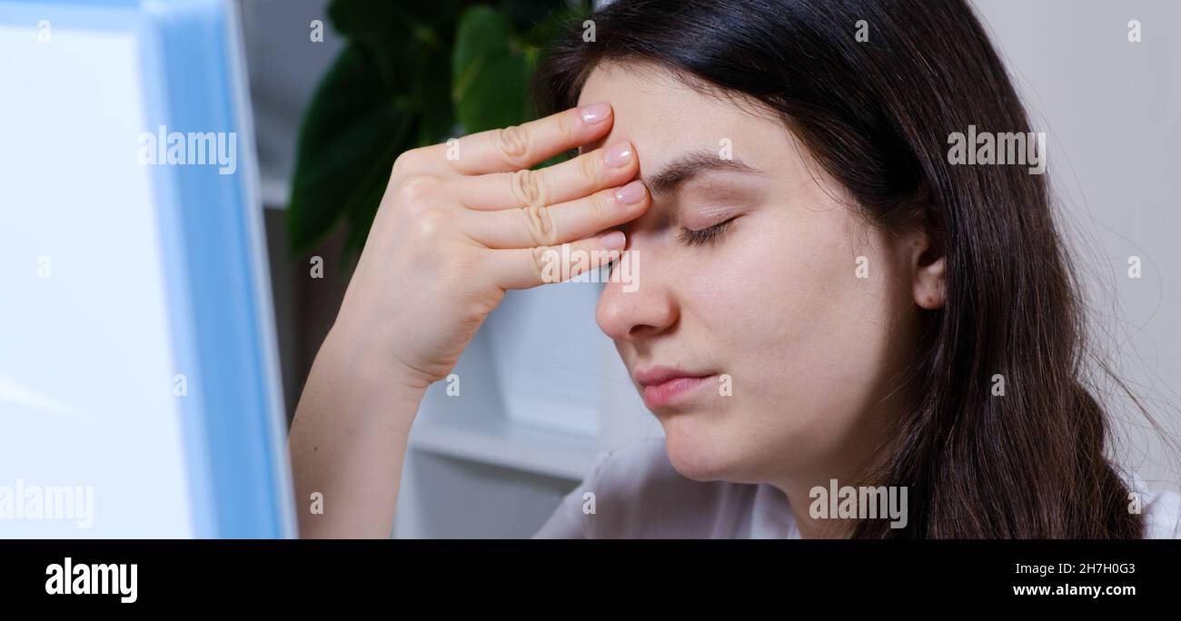 Una donna stanca con gli occhi chiusi tiene la mano sulla fronte mentre si siede di fronte a uno schermo del computer Foto Stock