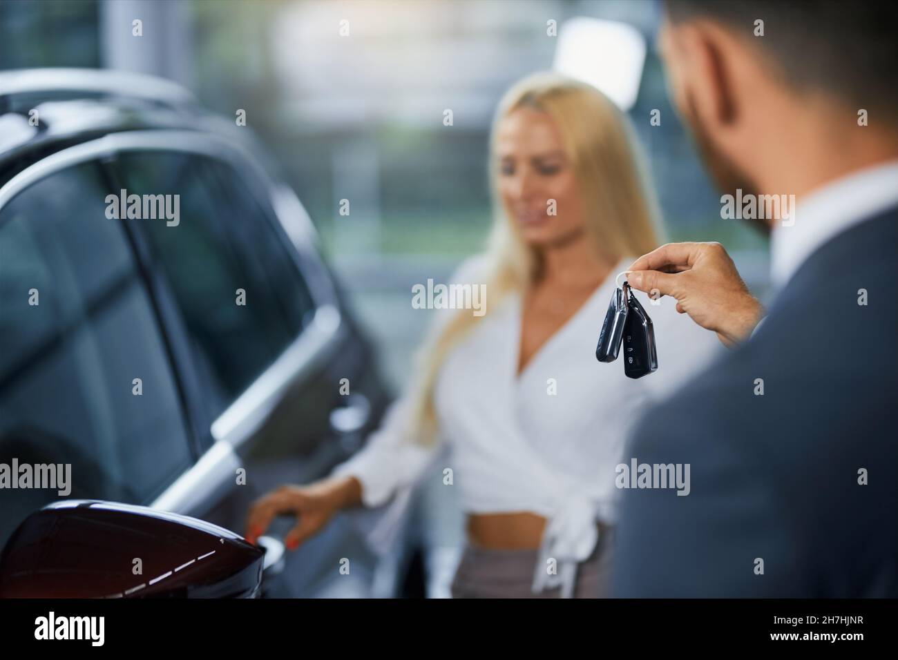Primo piano del concessionario di auto in tuta passando le chiavi al nuovo proprietario di auto, mentre in piedi insieme al salone di auto. Sfondo sfocato di donna caucasica in piedi vicino al suo trasporto di lusso. Foto Stock