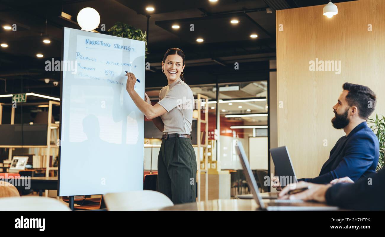 Giovane donna d'affari che presenta la sua strategia di marketing al suo team in un ambiente di lavoro moderno. Una donna felice manager sorridente mentre scriveva su un tabellone durante Foto Stock