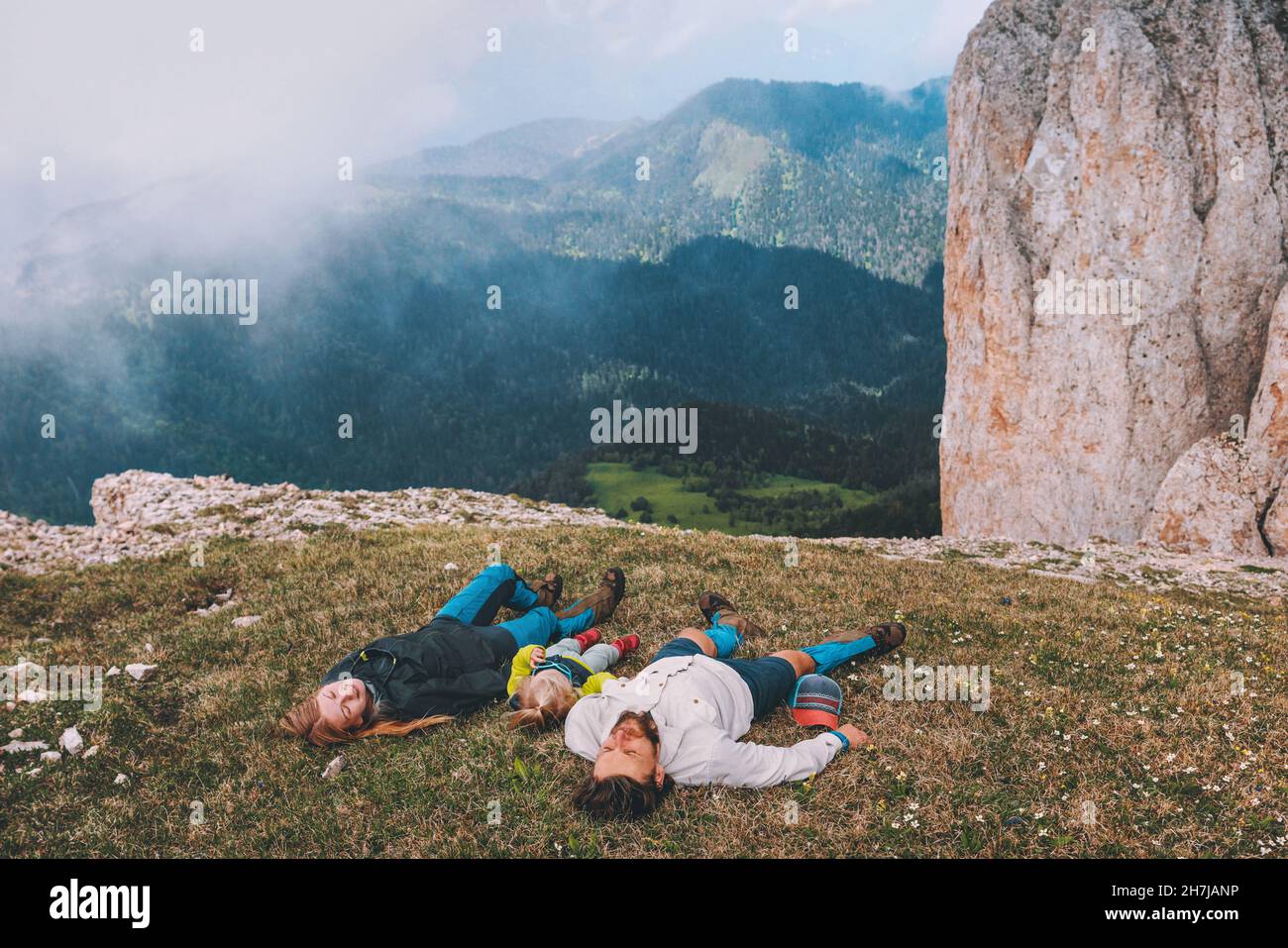 Famiglia che posa su erba in montagna madre padre con bambino insieme viaggio escursionismo all'aperto sano stile di vita persone rilassarsi godendo di vista delle nuvole Foto Stock