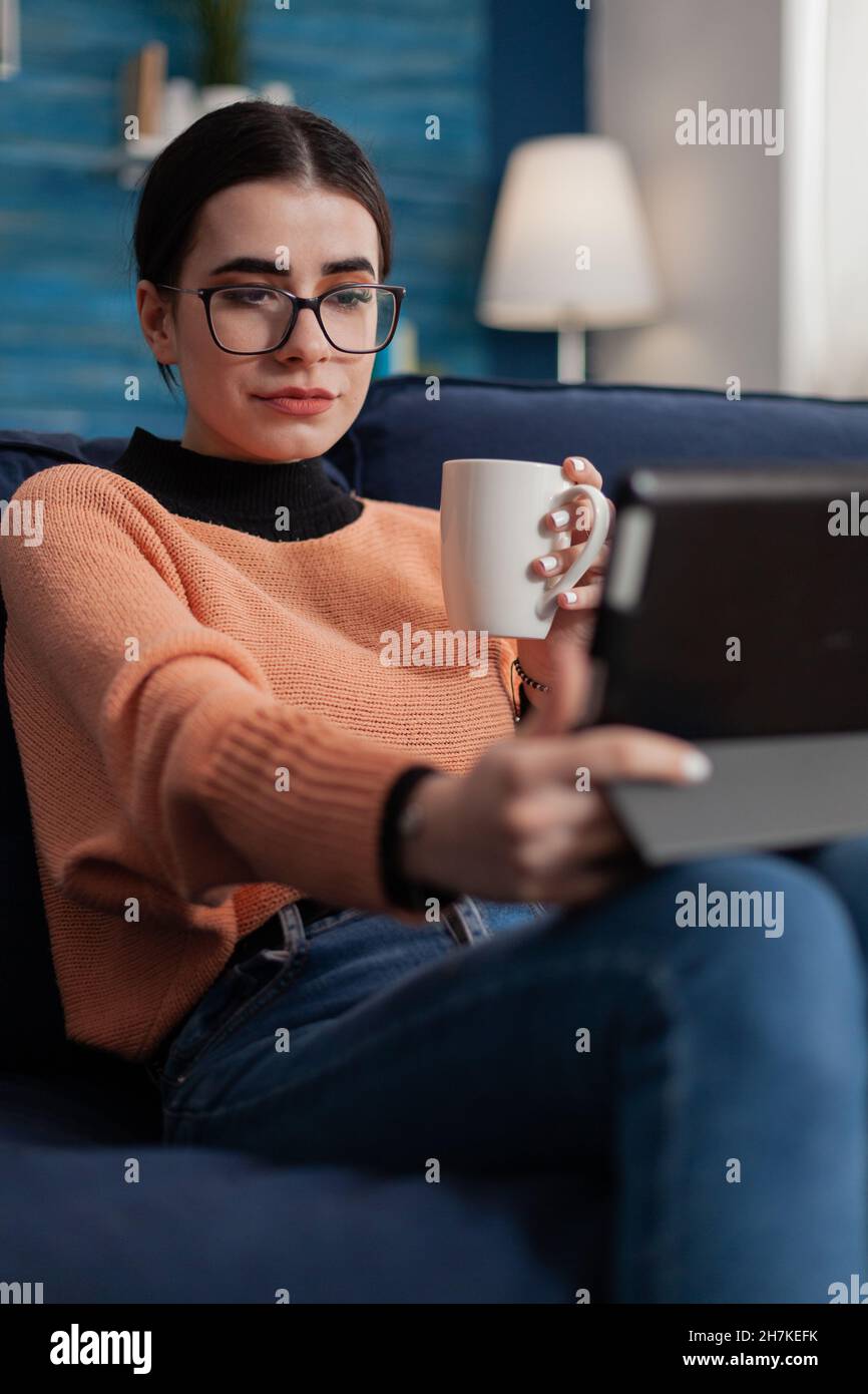 Studente con occhiali sul divano che tiene la tazza guardando il display del tablet. Influencer sul divano in videoconferenza mentre si beve caffè o tè. Creazione di contenuti per lo streaming di video su un dispositivo digitale. Foto Stock