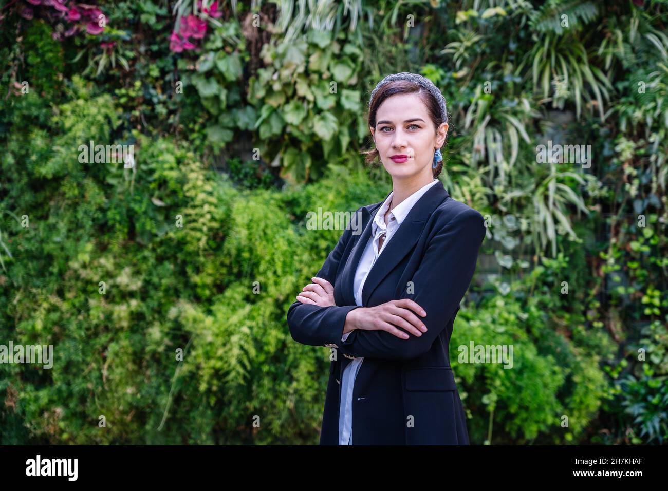Dipendente giovane donna assicurato con capelli scuri in abiti formali in piedi nel parco vicino a lussureggianti cespugli verdi con braccia incrociate e guardando la macchina fotografica Foto Stock
