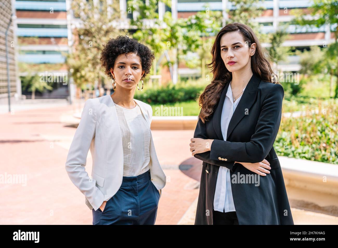 Giovani e fidati collaboratori di sesso femminile in abiti eleganti in piedi nel parco cittadino con braccia e mani ripiegate in tasca e guardando la fotocamera Foto Stock