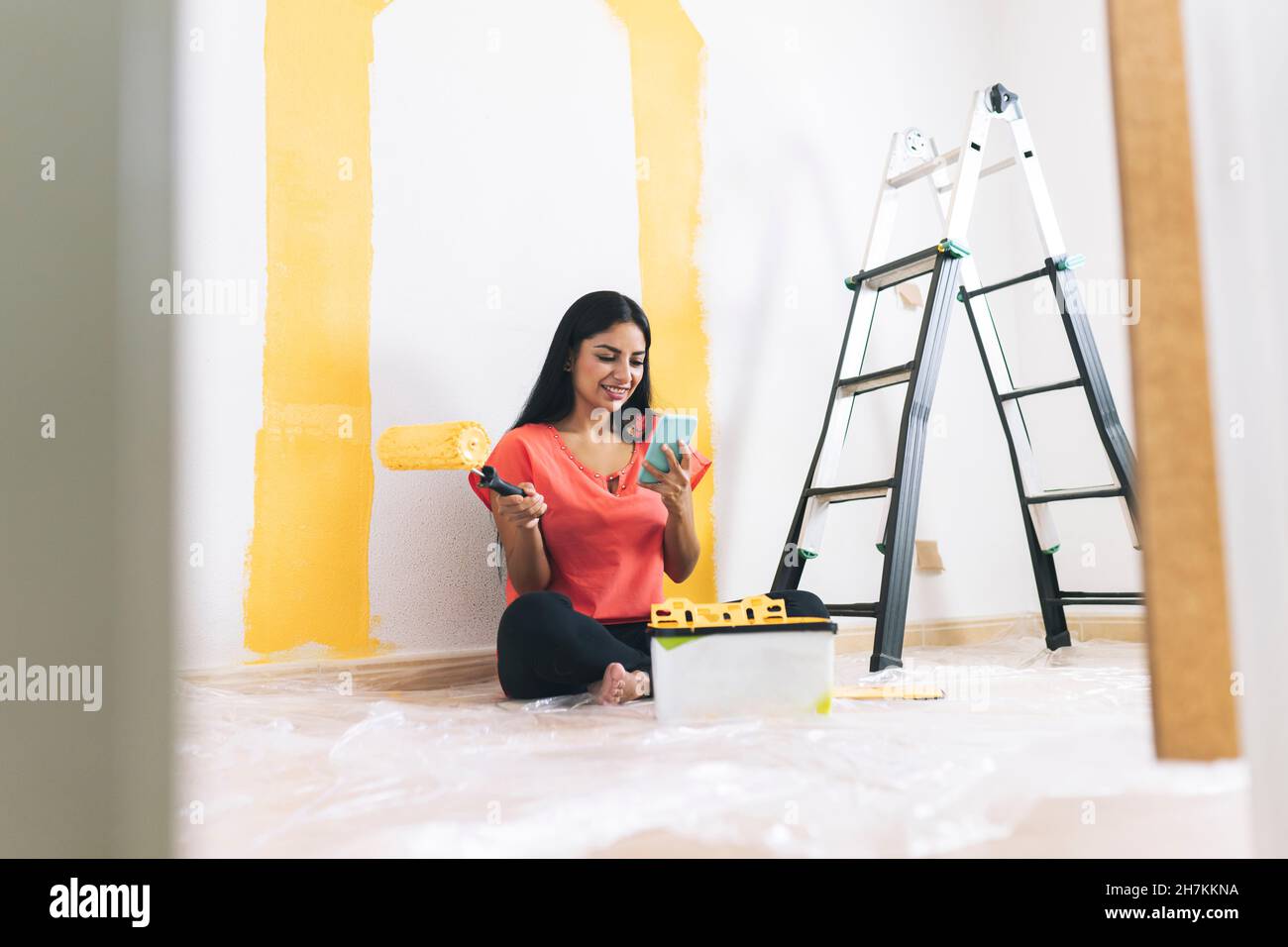 Giovane donna sorridente con rullo di vernice seduto a casa a scala Foto Stock
