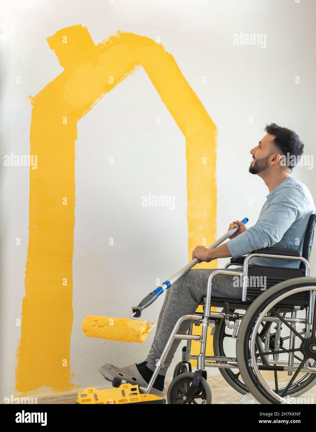 Uomo sorridente che tiene il rullo di vernice mentre si siede sulla sedia a rotelle nel soggiorno Foto Stock