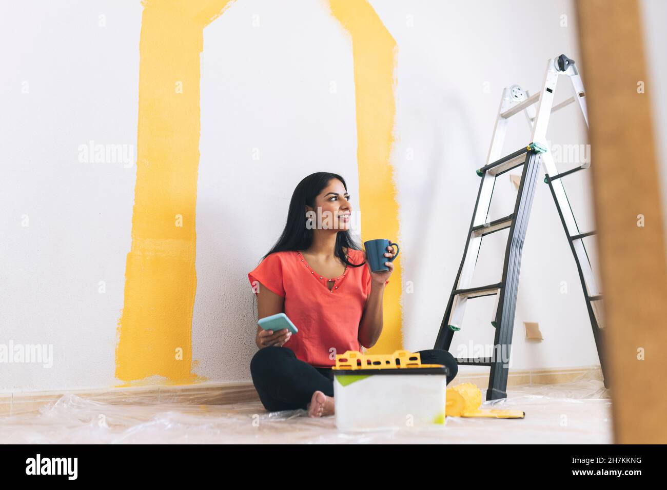 Giovane donna sorridente con rullo a vernice che usa lo smartphone in salotto Foto Stock