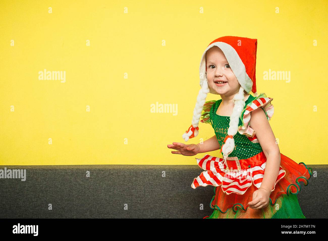 Ragazza in abito piccolo elfo di babbo natale galoppa in sala sul divano. Bambino in cappello Miss Babbo Natale con pigtail gioca la notte di Natale in attesa di regali Foto Stock