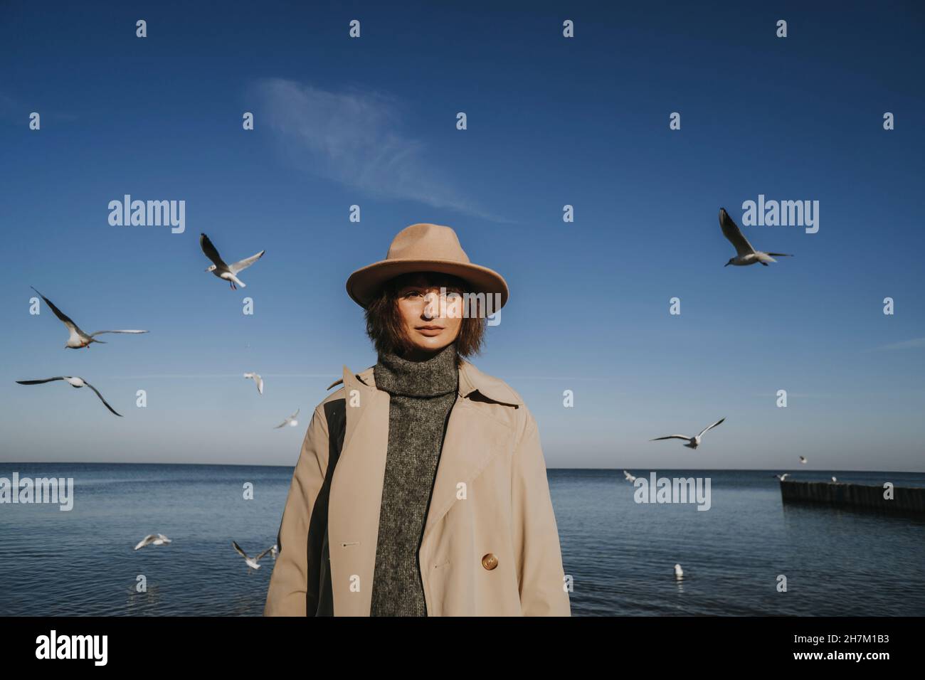 Donna in soprabito e cappello in piedi davanti ai gabbiani che volano sul mare Foto Stock