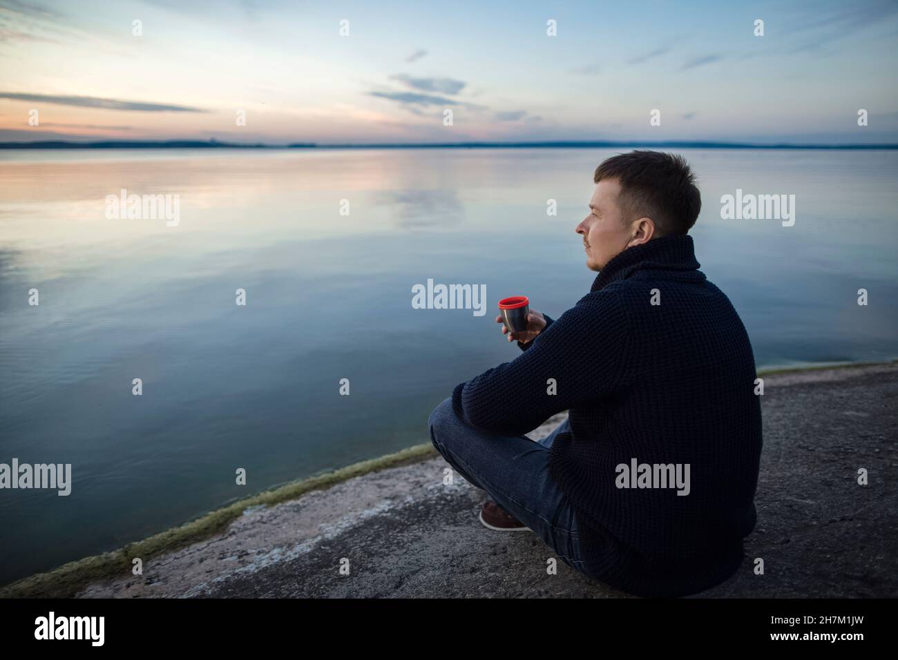 Uomo che beve il tè seduto sulla roccia vicino al lago al tramonto Foto Stock