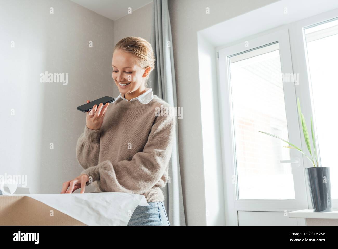 Donna sorridente con un piano di invio di messaggi vocali tramite telefono cellulare a casa Foto Stock