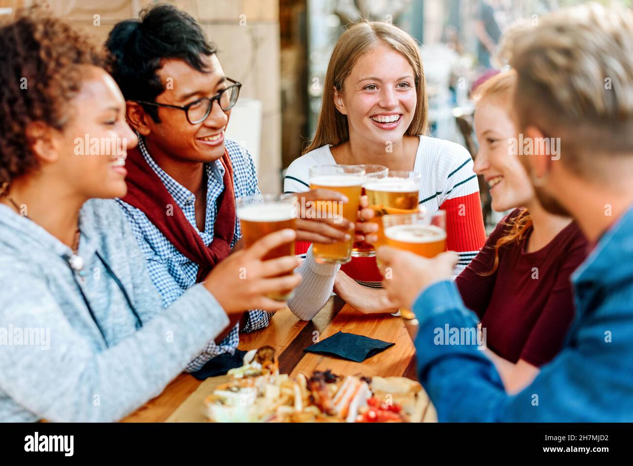 Gruppo di giovani amici diversi godendo di una birra insieme alzando i loro bicchieri in un brindisi con fuoco a una giovane donna bionda sorridente Foto Stock