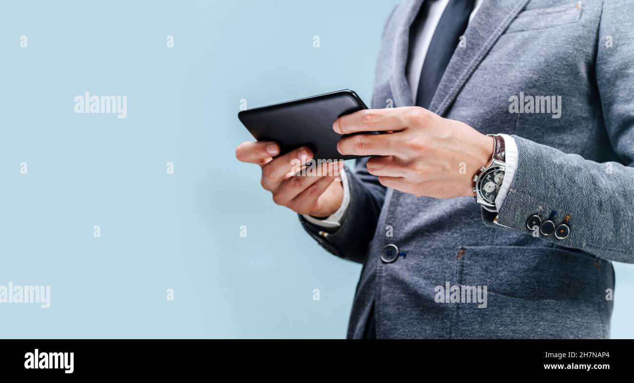 Un uomo d'affari giovane in una giacca e cravatta con un orologio in mano tiene un telefono, mani da vicino su uno sfondo blu Foto Stock