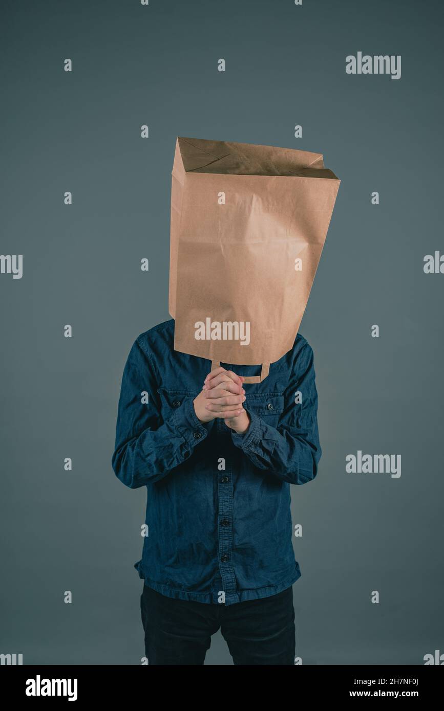 Giovane uomo con un sacchetto di carta sulla testa, le mani si piegano per pregare, concetto di speranza, sfondo grigio scuro Foto Stock