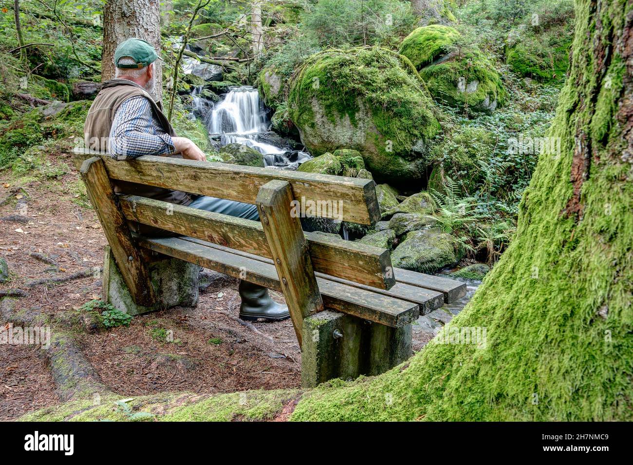 Uno zaino in spalla siede su una panca di legno solitario nella foresta e guarda la piccola cascata. Foto Stock