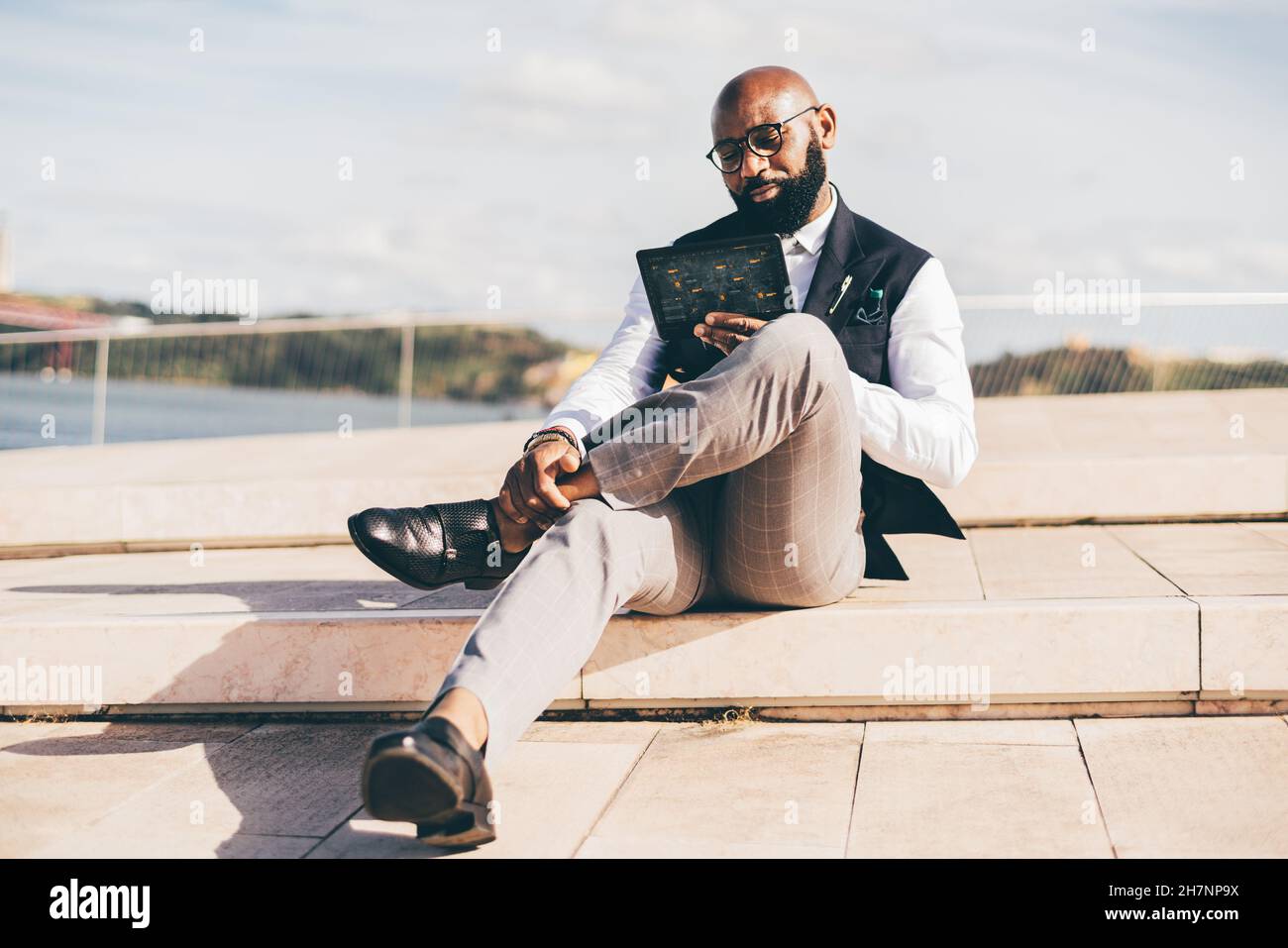 Un bel calvo bearded uomo nero imprenditore in occhiali e un vestito formale con un gilet, è seduta all'aperto e interagire con un tabl futuristico Foto Stock