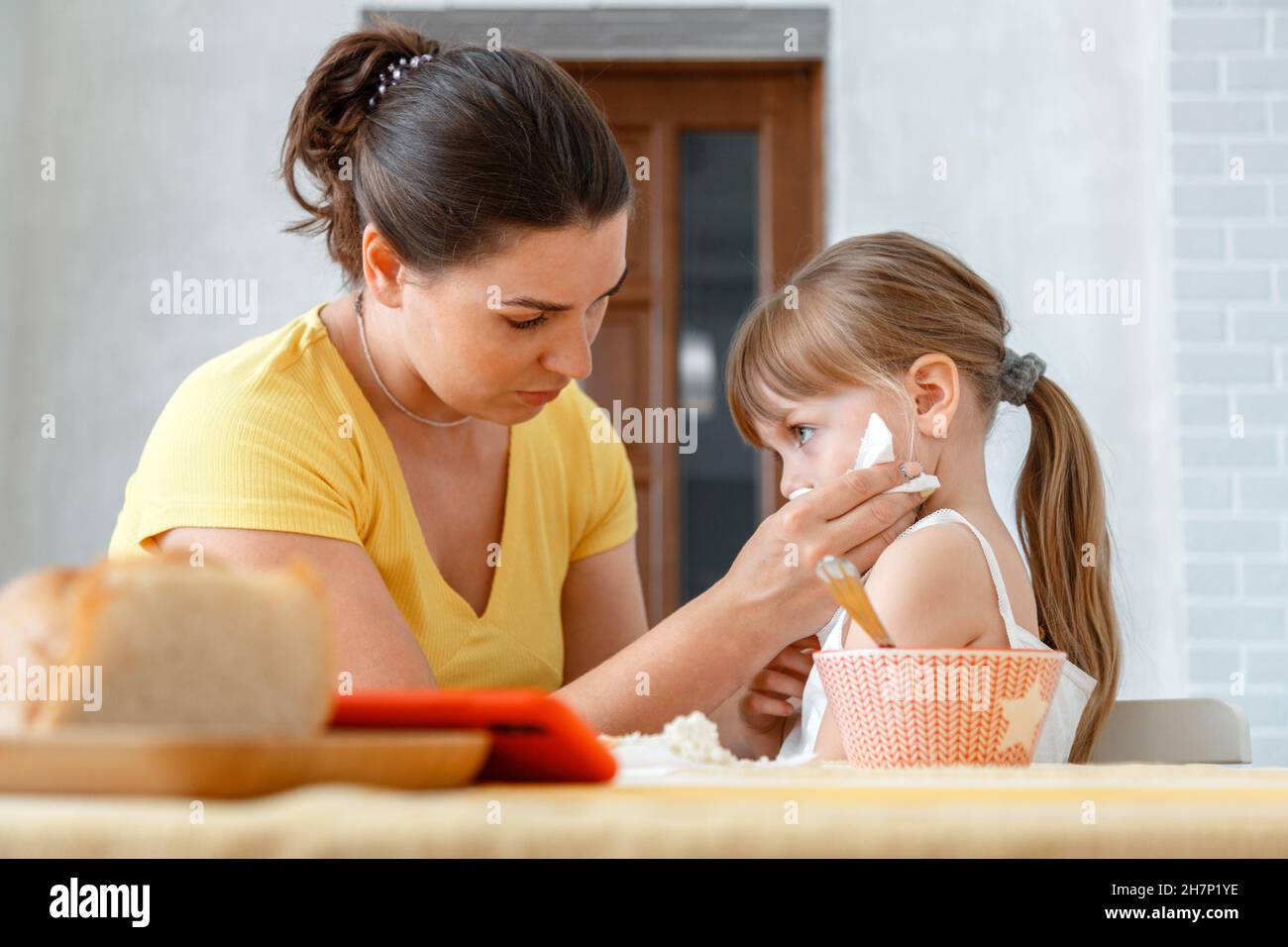 La madre nutre la figlia con il cibo dal cucchiaio e la pulisce con il tovagliolo. Cura materna del bambino bambino ragazza reale lifestyle pranzo di delusa Foto Stock