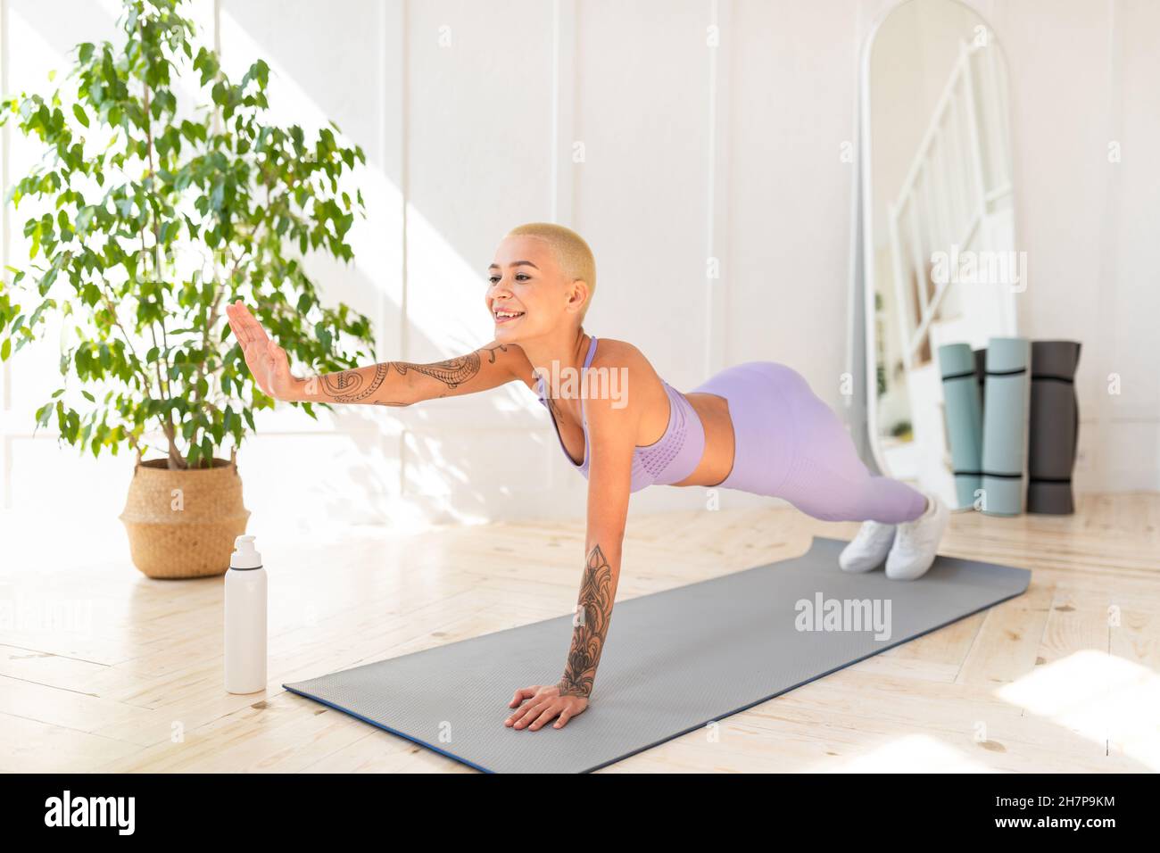 Una giovane donna sportiva che si allena a casa, fa una tavola di yoga e solleva una mano, esercitandosi sul tappeto di yoga in una stanza luminosa Foto Stock