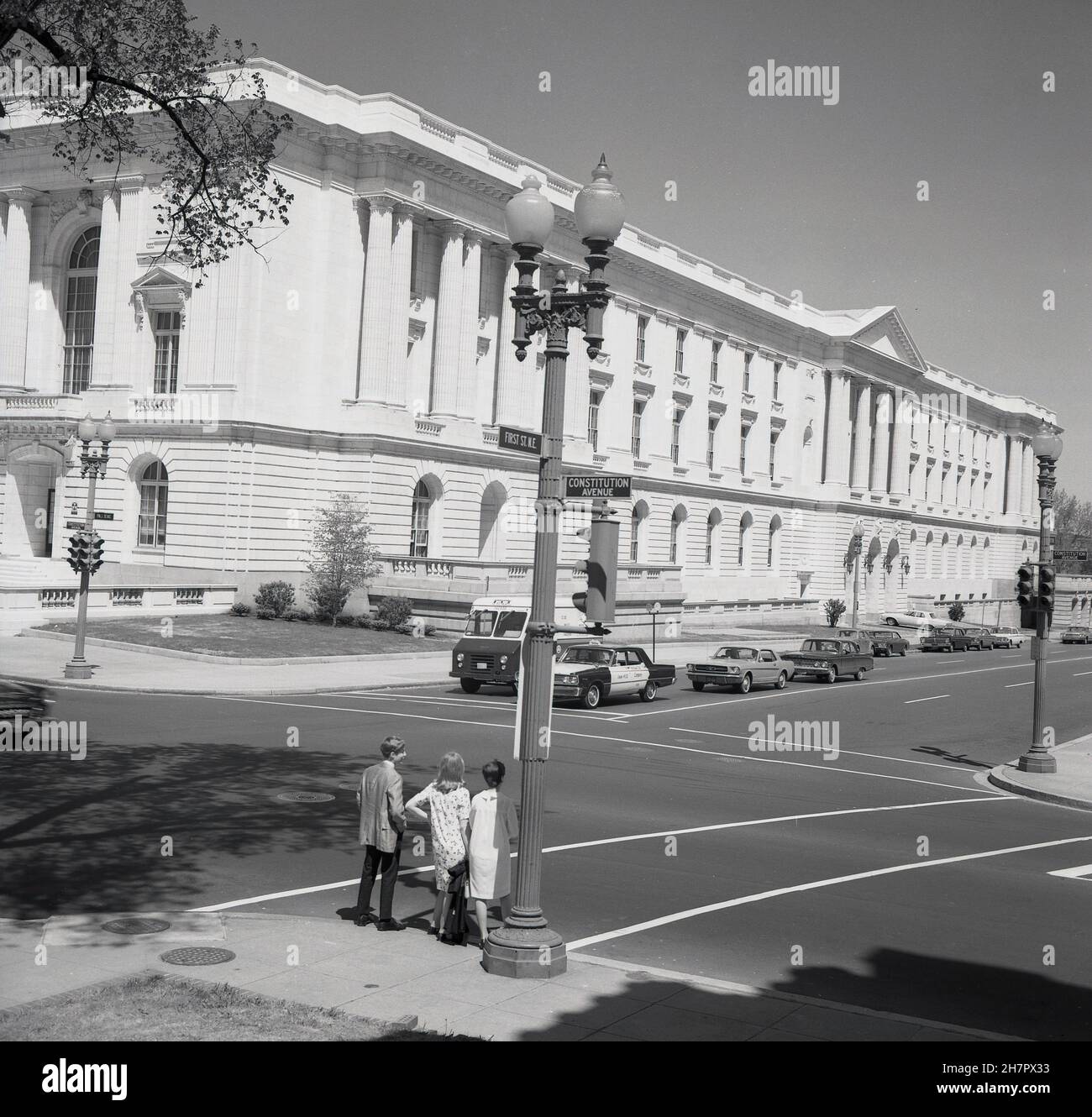 1960s, storico, un giovane e due giovani donne in attesa di attraversare Constitution Avenue a Washington DC, USA, la capitale del paese. All'incrocio opposto, le automobili dell'epoca in attesa di attraversare la strada, un taxi giallo, e un camion postale degli Stati Uniti. I principali dipartimenti governativi degli Stati Uniti si trovano in molti dei grandi edifici colonnati neoclassici della strada. Originalmente noto semplicemente come B St. NW, quando l'area fu risviluppata alla fine del 1930s, fu suggerito di rinominarla Constitution Avenue in onore del terzo presidente americano Thomas Jefferson e fondatore Padre, e questo ebbe luogo nel 1943. Foto Stock