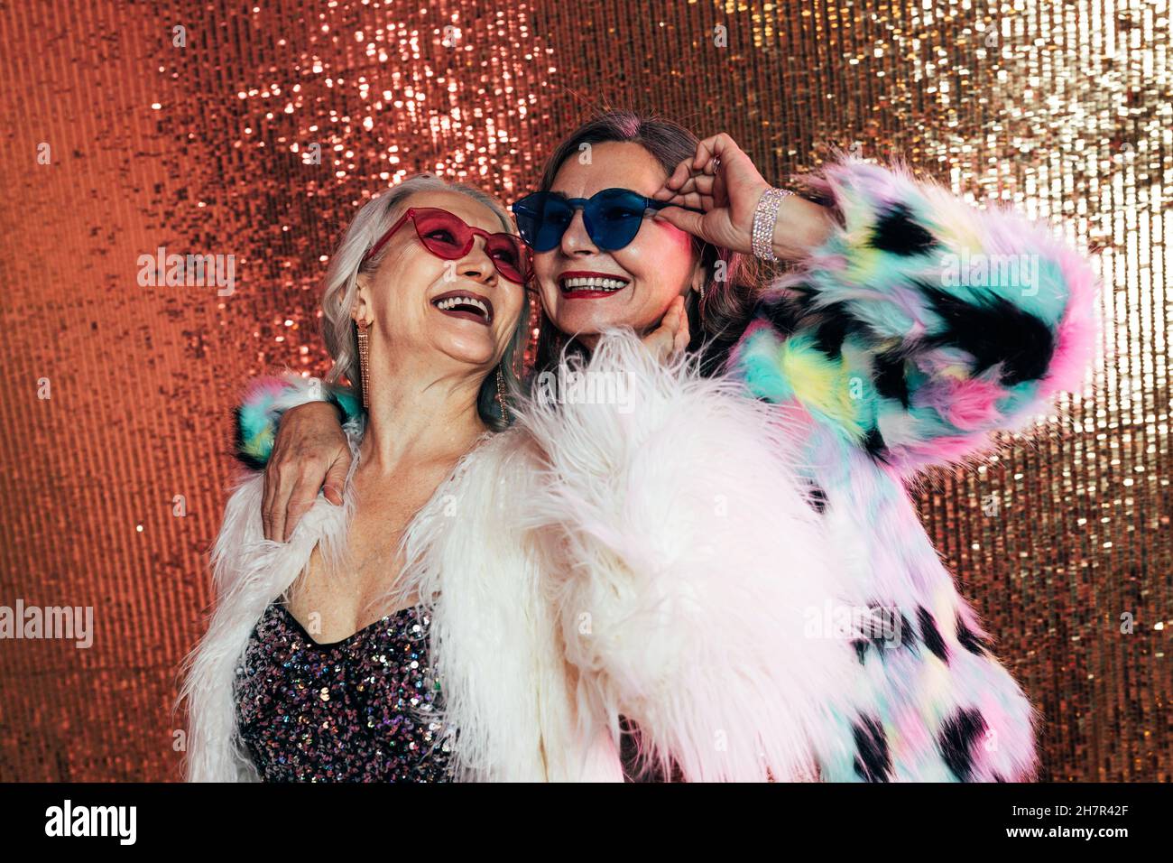 Due donne che indossano occhiali e cappotti in pelliccia. Le donne anziane ridono sullo sfondo glitterato. Foto Stock