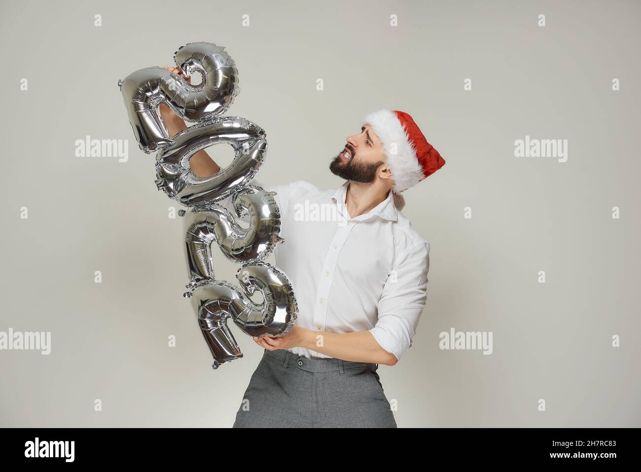 Un uomo trattante in un velluto rosso Santa Hat sta spingendo palloncini d'argento a forma di 2022 a sinistra verticale. Un ragazzo con una barba a una festa di Capodanno. Foto Stock